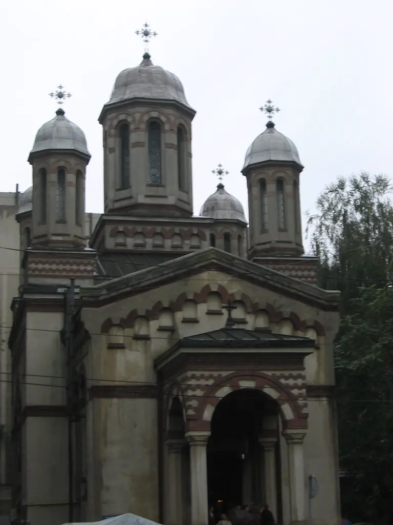 Fachada de uma igreja ortodoxa romena com quatro cúpulas cinzentas encimadas por cruzes, arcos de tijolo aparente e um pórtico na entrada.